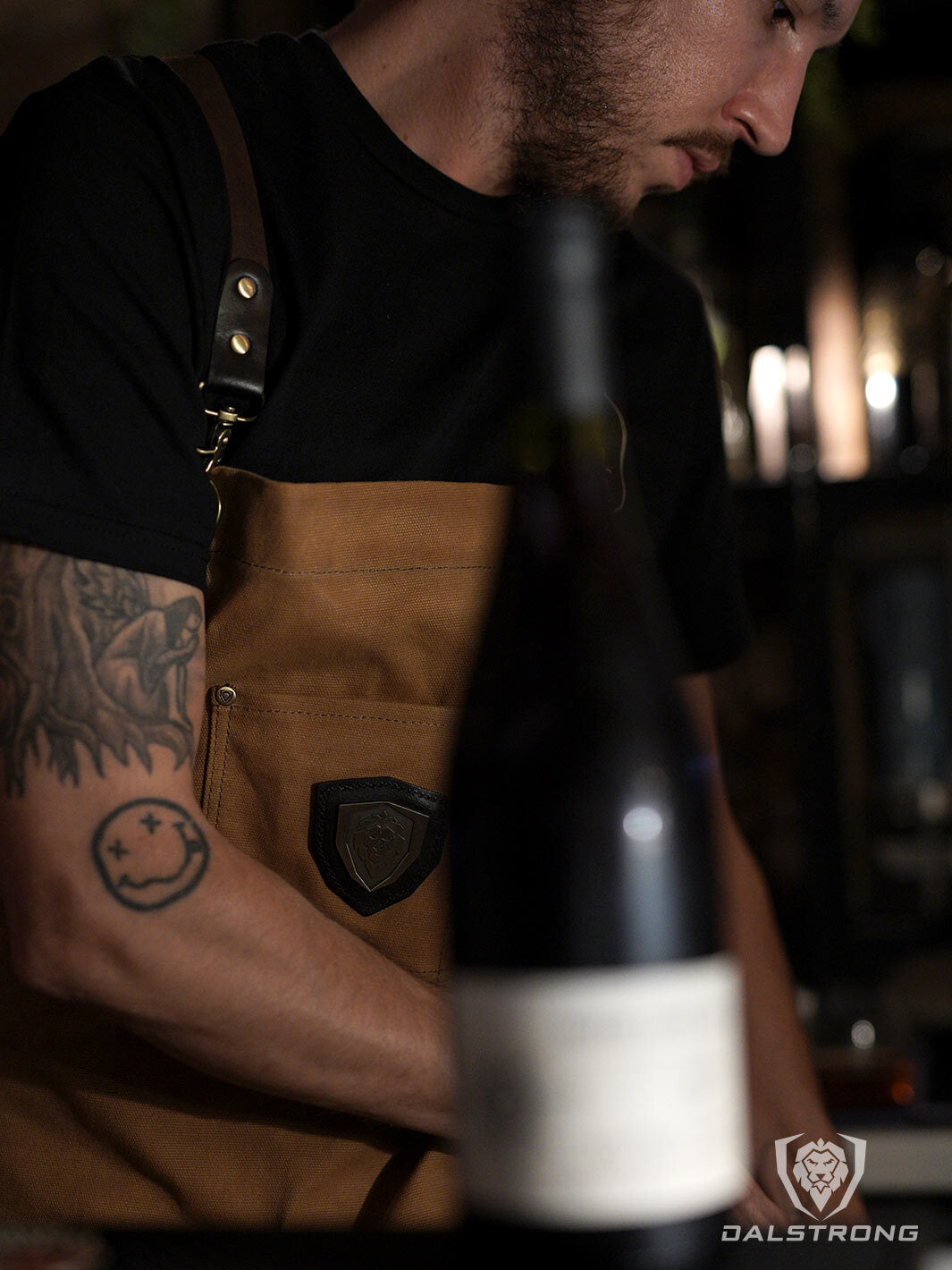 A man with tattoo wearing the Dalstrong heavy-duty waxed canvas professional chef apron brown dessrt drifter.