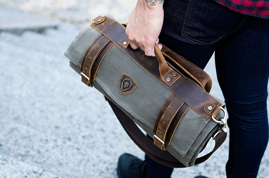 A man with pants is holding the Dalstrong 12oz heavy-duty canvas and leather olive nomad knife roll.