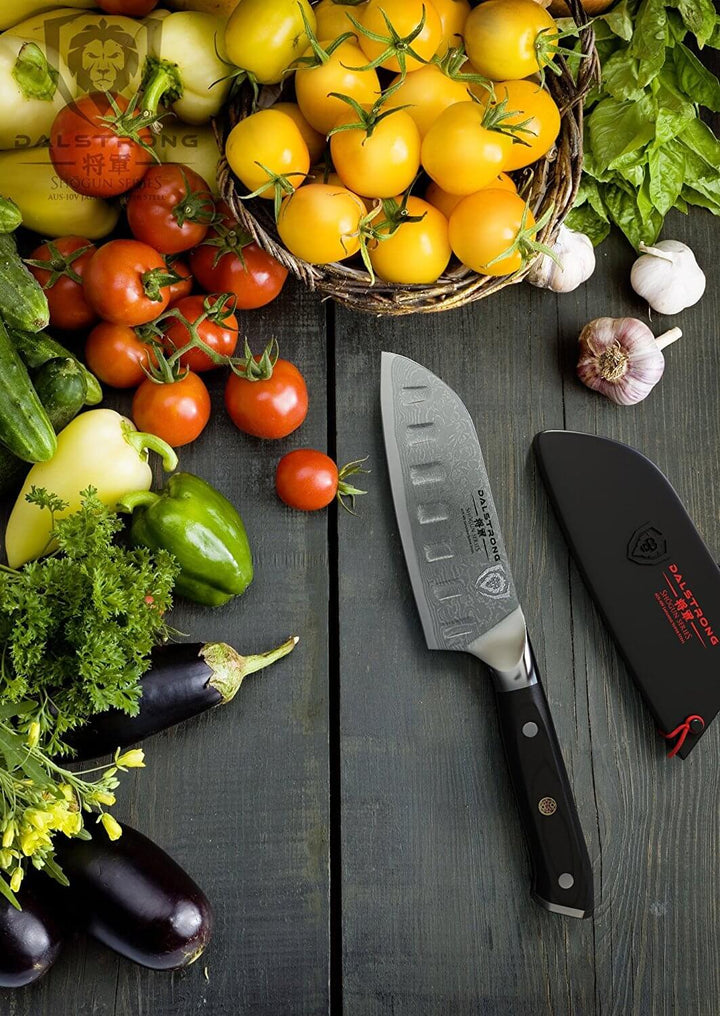 Dalstrong shogun series 5 inch santoku knife with black handle and different kinds of vegetables on the side.