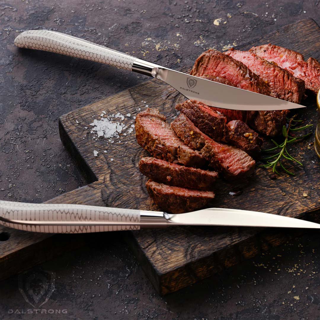 Dalstrong frost fire series steak knife set with white honeycomb handle and slices of steak on a cutting board.
