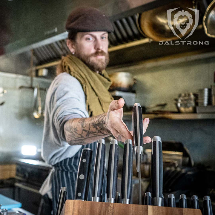 A chef grabbing a knife from the dalstrong gladiator series 18 piece knife set with black handles and block.