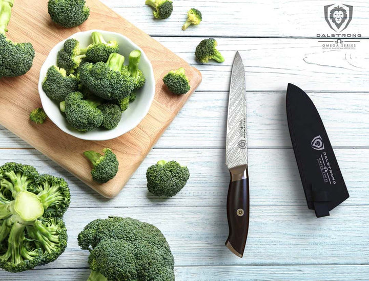 Dalstrong omega series 5.5 inch utility knife with black sheath beside some broccoli on a cutting board.