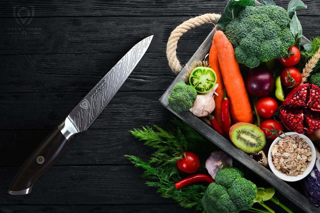 Dalstrong omega series 5.5 inch utility knife with black sheath beside some vegetables on a wooden container.