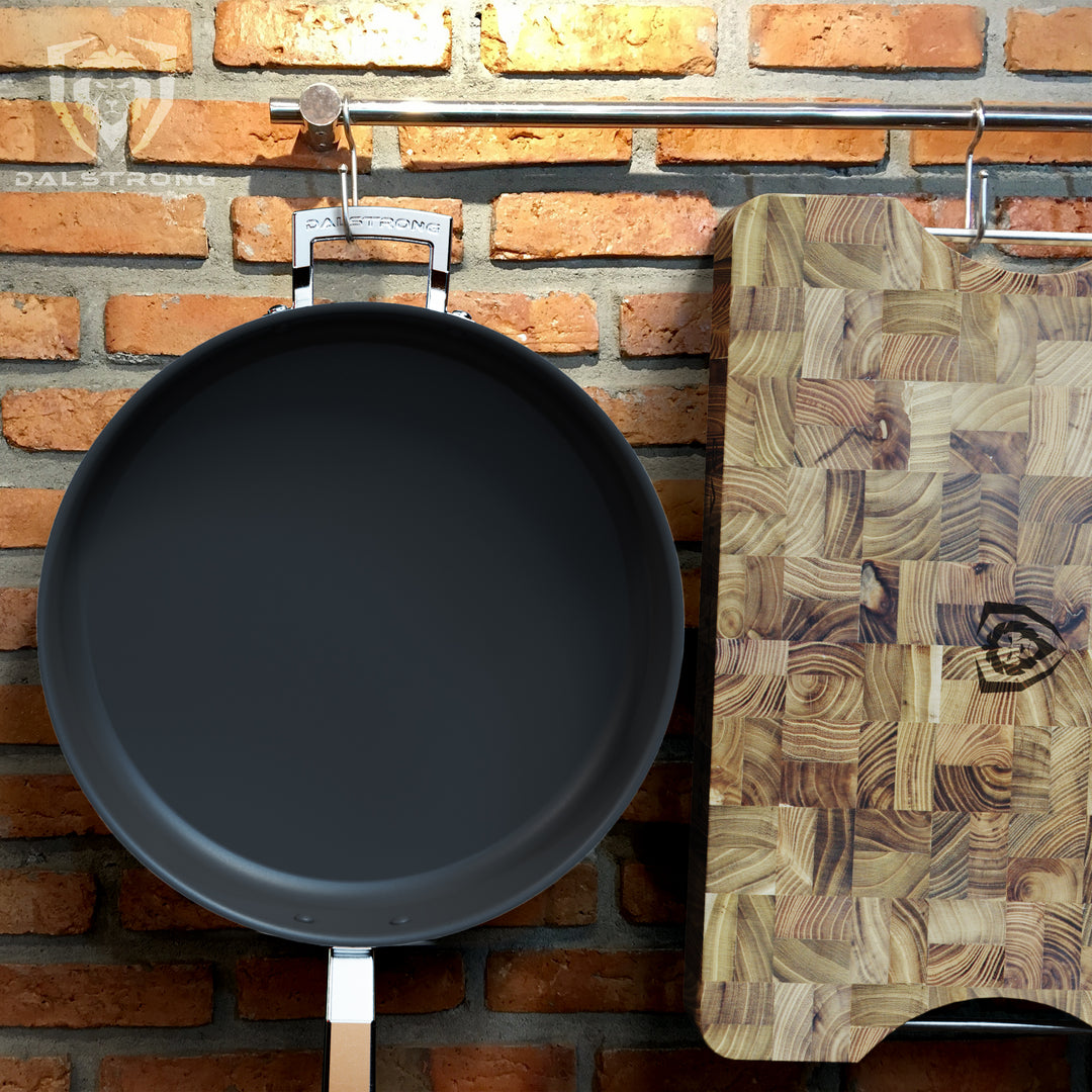Dalstrong Frying Pan and wooden cutting board hanging on a silver kitchen rod against a red bricked wall