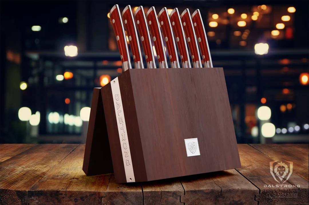Dalstrong gladiator series 8 piece steak knife set with red handles inside of it's handcrafted storage block on top of a wooden table.