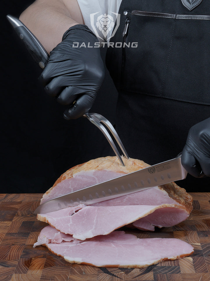 Dalstrong gladiator series 9 inch carving knife and fork set with a sliced ham on a dalstrong wooden board.