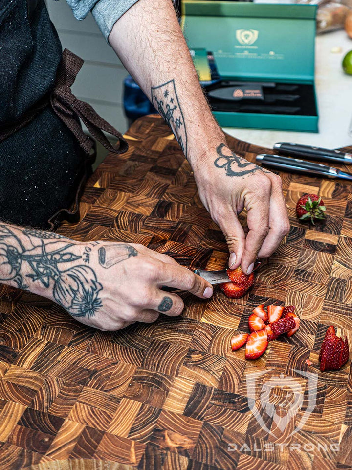 A hand with tattoo holding the dalstrong gladiator series 3 piece paring knife set with slices of strawberry on a dalstrong wooden board.