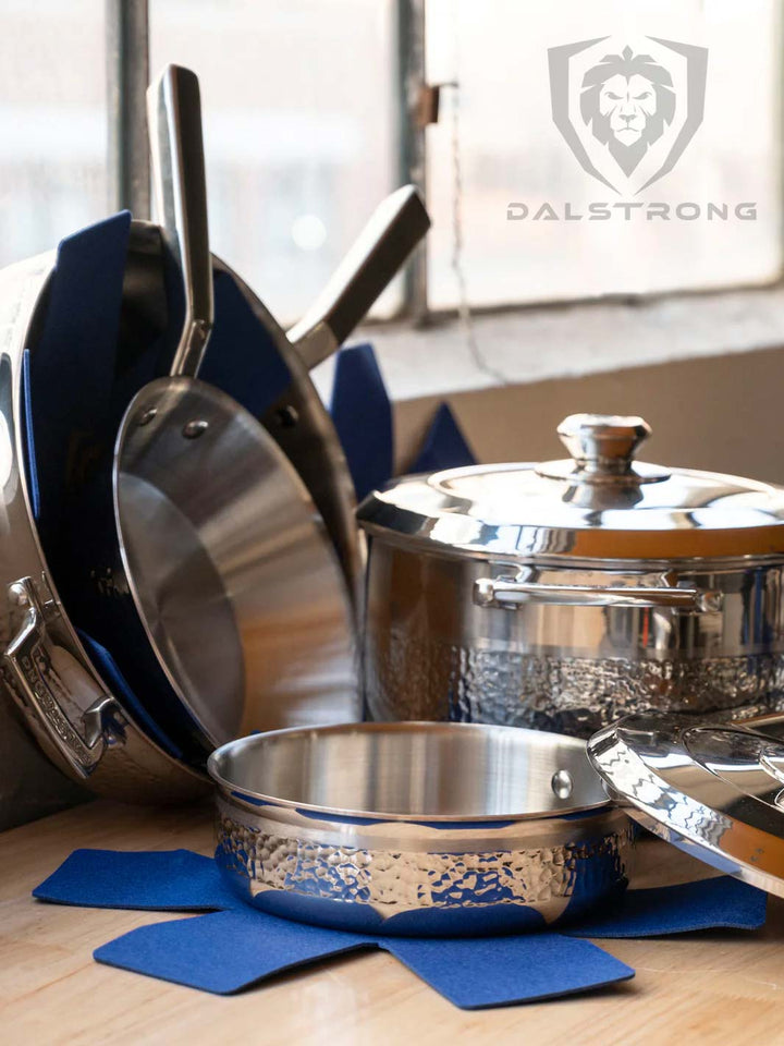 Dalstrong avalon series 12 piece cookware set silver on a kitchen table beside a window.