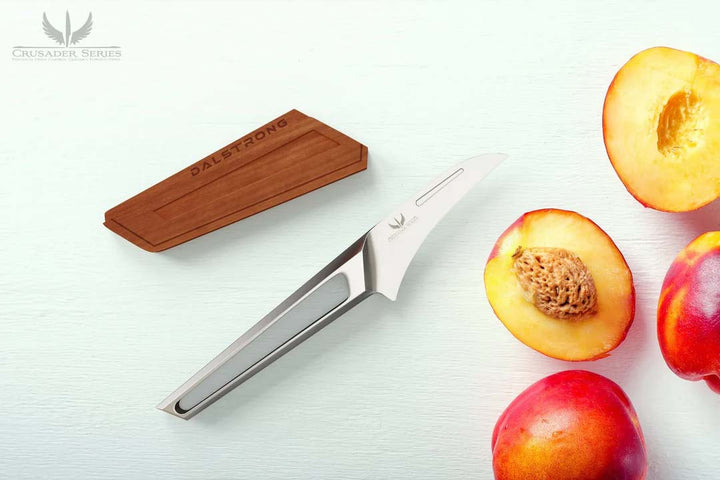Dalstrong crusader series 3 inch bird's beak paring knife with german steel handle and wooden sheath beside slices of peach.