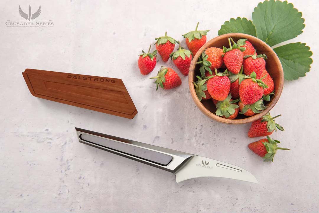 Dalstrong crusader series 3 inch bird's beak paring knife with german steel handle and wooden sheath beside some strawberries.