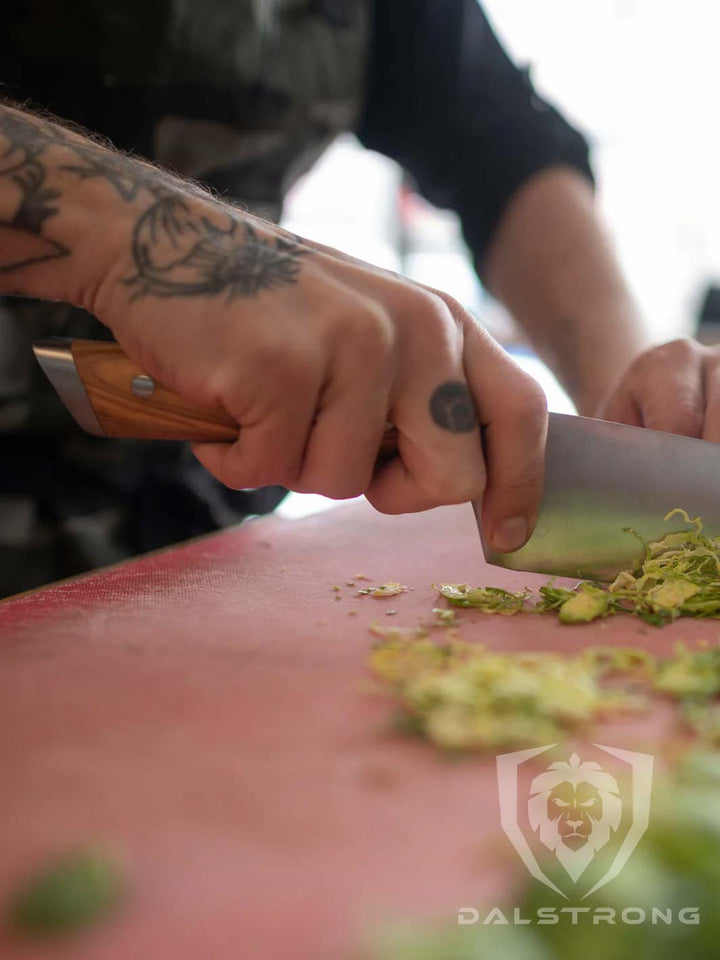 A man's hand with tattoo holding the dalstrong gladiator series 8 inch chef knife with olive wood handle and chopped brussel sprouts.