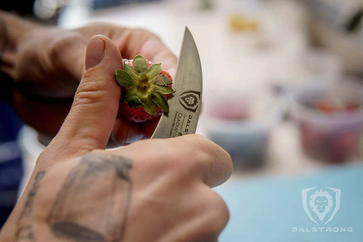 A man's hand with tattoo slicing a strawberry with dalstrong gladiator series 2.7 inch bird's beak paring knife.