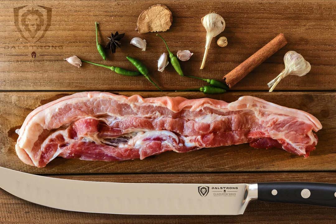 Dalstrong gladiator 10 inch butcher knife with black handle and a slab of pork belly on a wooden table.