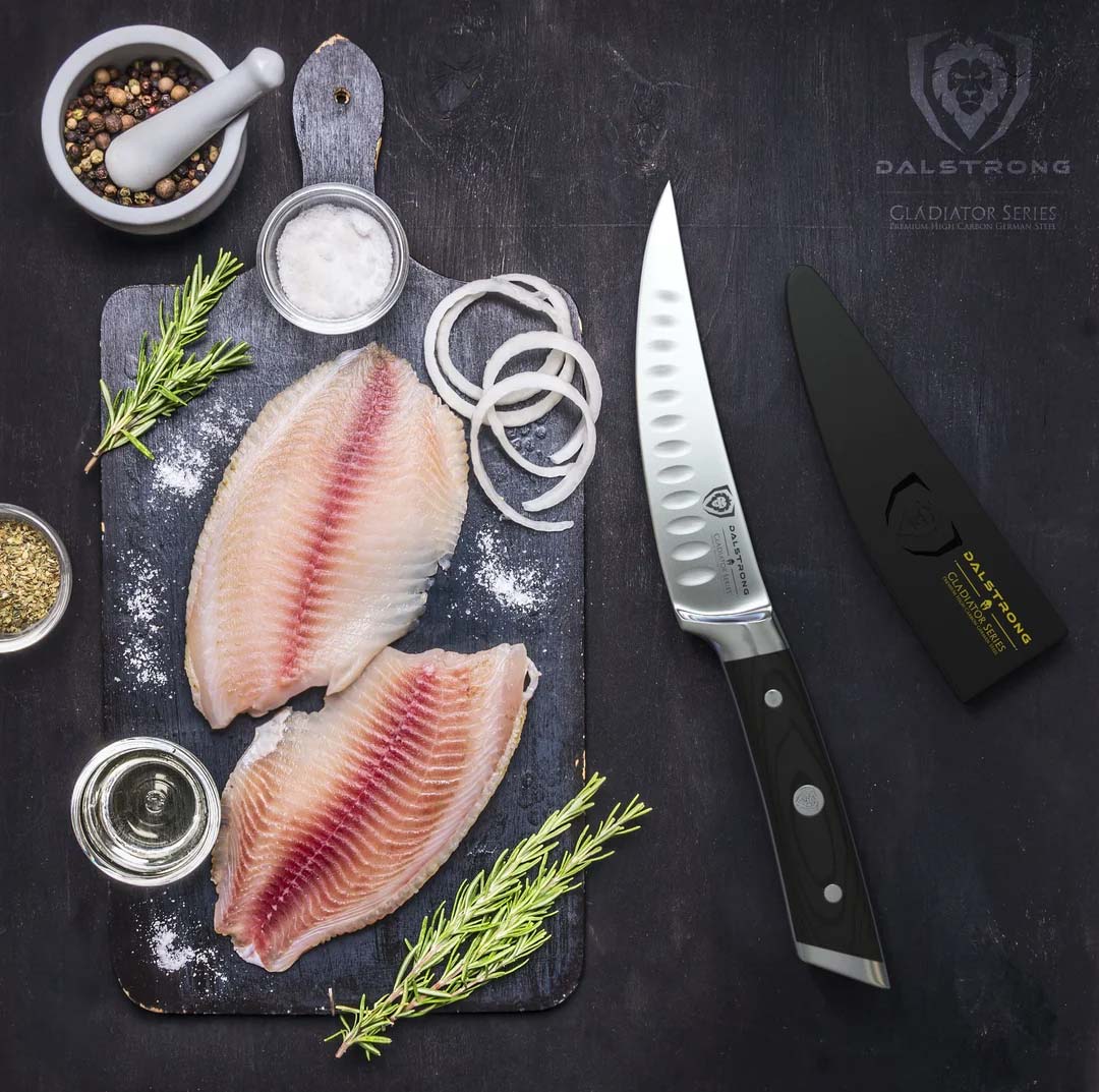 Dalstrong gladiator series 6 inch curved fillet knife with black handle and sheath beside two fillets of fish on a wooden board.