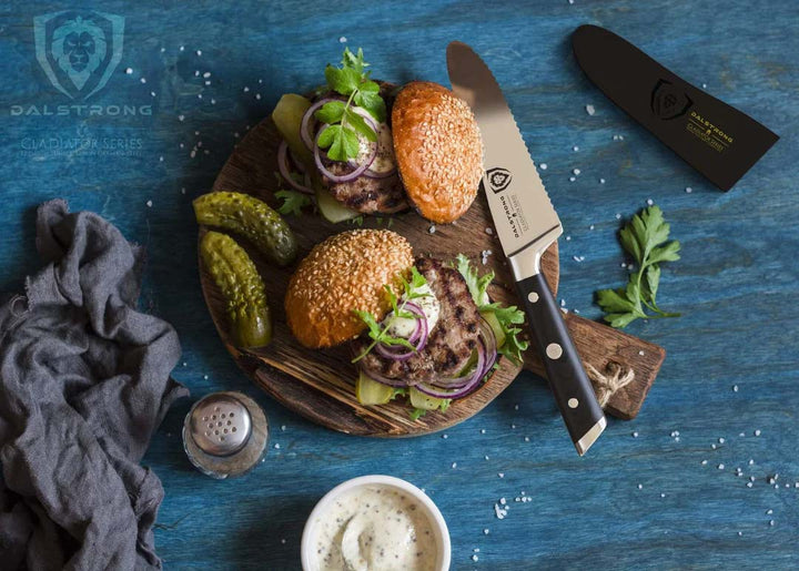 Dalstrong gladiator series 6 inch serrated sandwich knife with black handle and two hamburgers on a wooden cutting board.