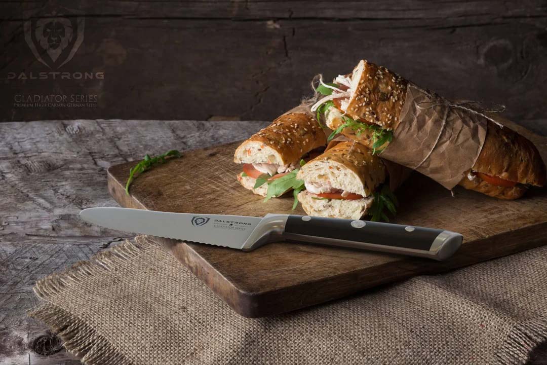 Dalstrong gladiator series 6 inch serrated sandwich knife with black handle and three sancwiches on a wooden cutting board.
