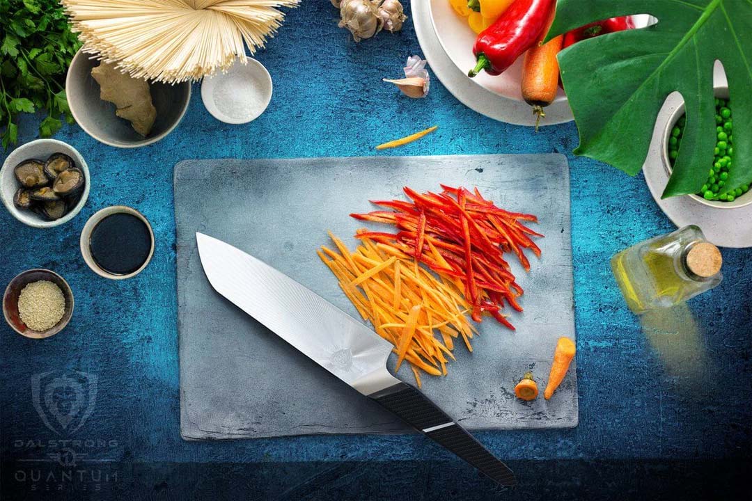 Dalstrong quantum 1 series 7 inch santoku knife with dragon skin handle beside thin slices of bell peppers and carrots.