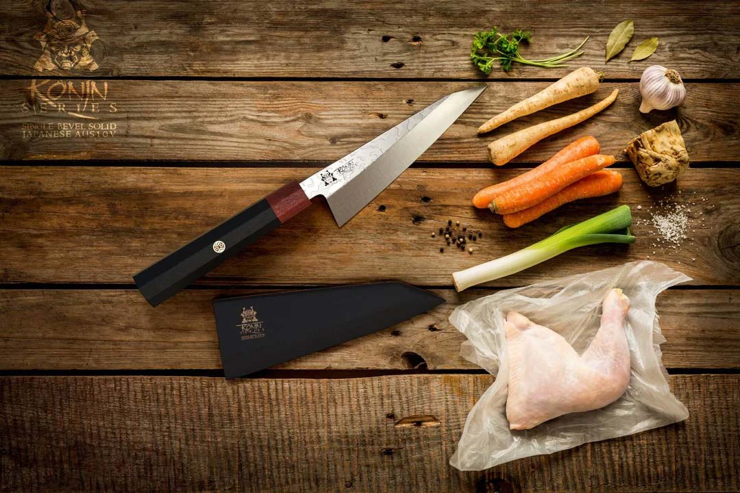 Dalstrong ronin series 5.5 inch honesuki knife with black handle and sheath beside a slice of chicken meat and vegetables.