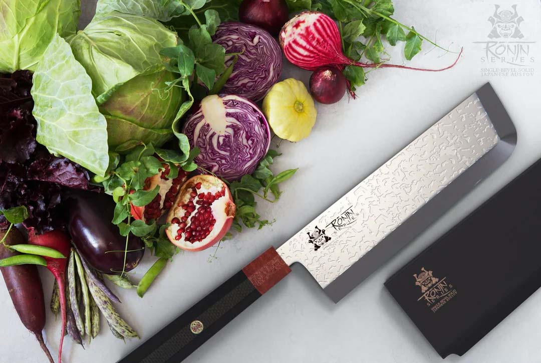 Dalstrong ronin series 7 inch usuba knife with black handle and sheath beside some cabbage and eggplant.