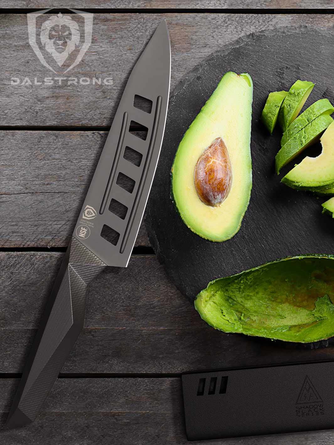 Dalstrong shadow black series 7 inch vegetable knife and black sheath beside slices of green avocado on a wooden board.
