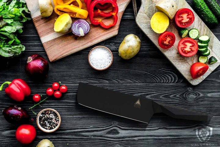 Dalstrong shadow black series 7 inch nakiri knife with black sheath beside some tomato, potatoes and bell pepper cut in half.
