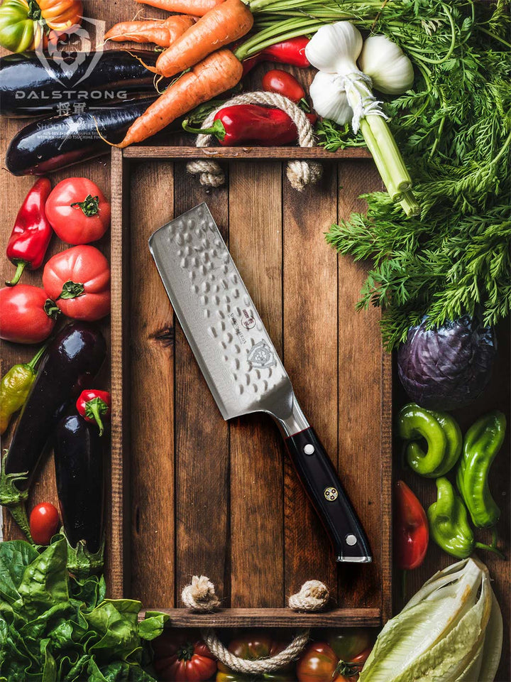 Dalstrong shogun series 6 inch nakiri knife with black handle in a wooden board surrounded by different kinds of vegetables.