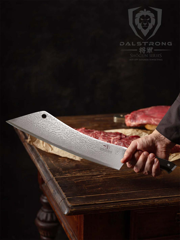 A hand holding the Dalstrong shogun series 12 inch crixus cleaver knife with black handle and a meat on a wooden table.