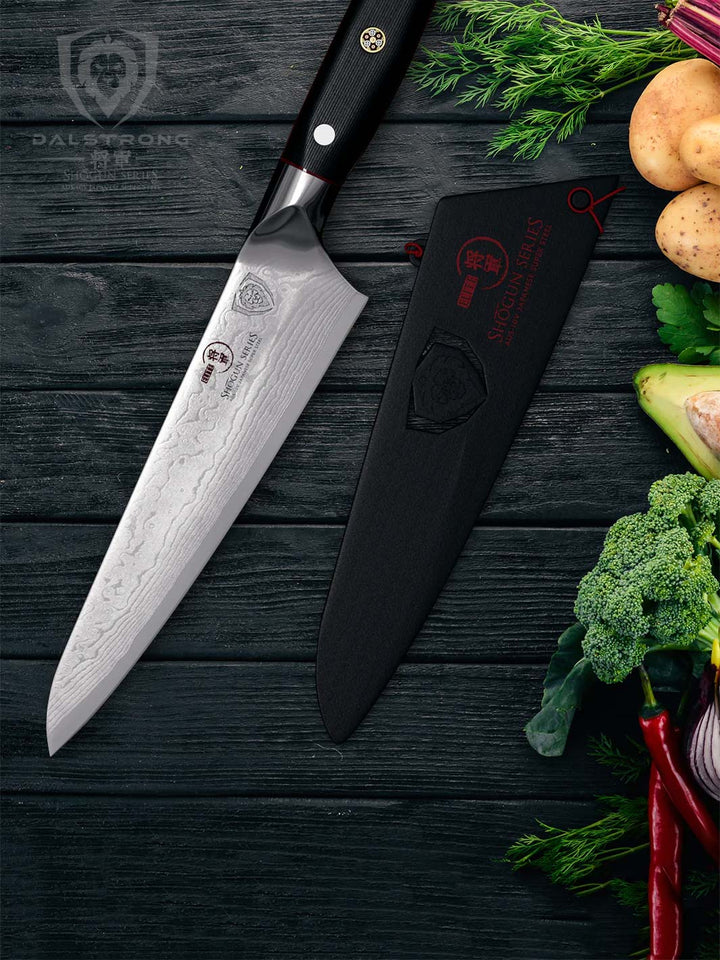 Dalstrong shogun series 7 inch chef knife with black handle and sheath beside different kinds of vegetables.