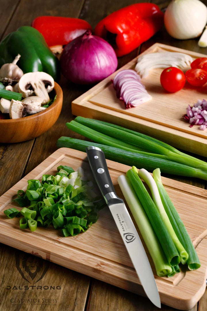 Dalstrong gladiator series 6 inch utility knife with black handle and slices of scallions on a wooden cutting board.