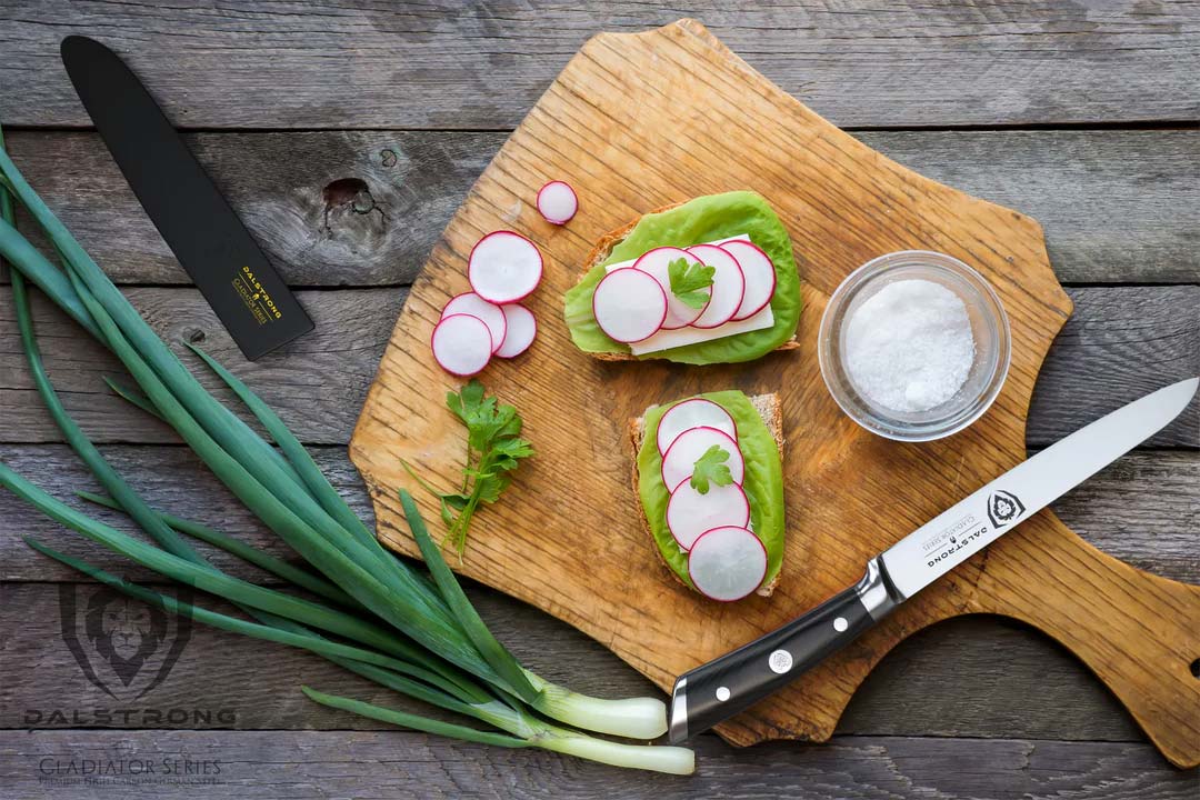 Dalstrong gladiator series 6 inch utility knife with black handle and slices of radish on a wooden cutting board.