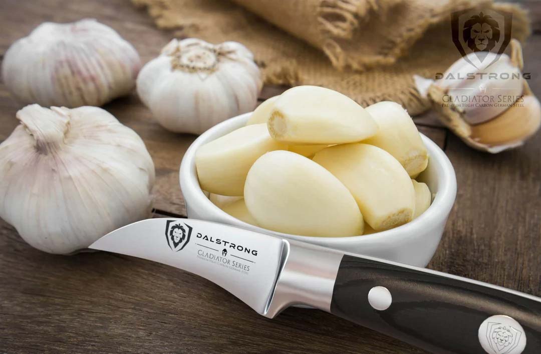 Dalstrong gladiator series 2.7 inch bird's beak paring knife with black handle and peeled garlic inside a cup.