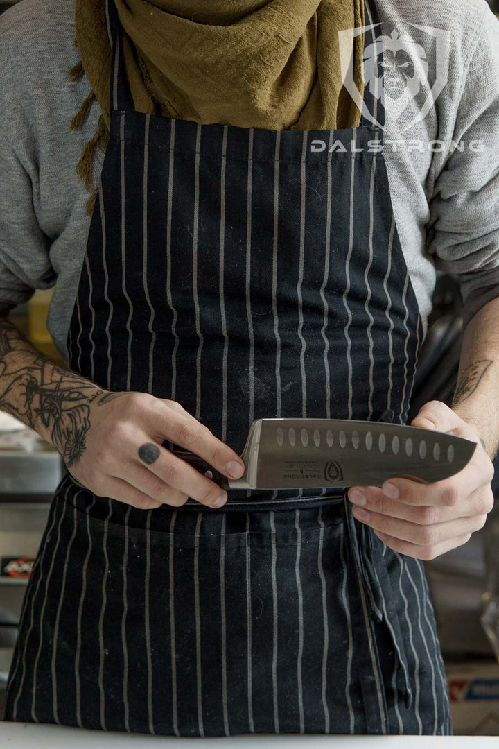 A man wearing an apron holding the dalstrong gladiator series 7 inch santoku knife with black handle.