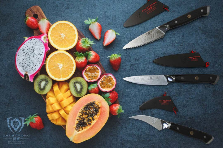 Dalstrong shogun series 3 piece paring knife set with black handles and sheath beside different kinds of fruits on a wooden board.